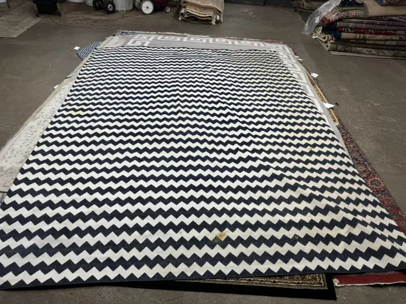 A black and white chevron rug laid out on a work floor, rolled rugs behind it